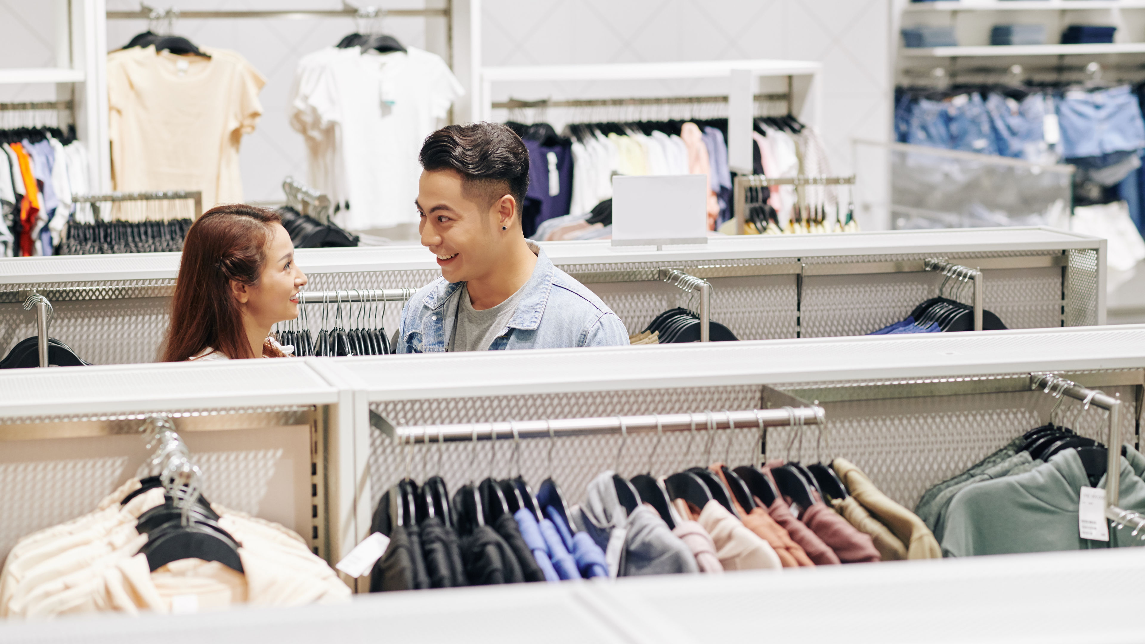Smiling couple shopping for clothes in a store, browsing through clothing racks.