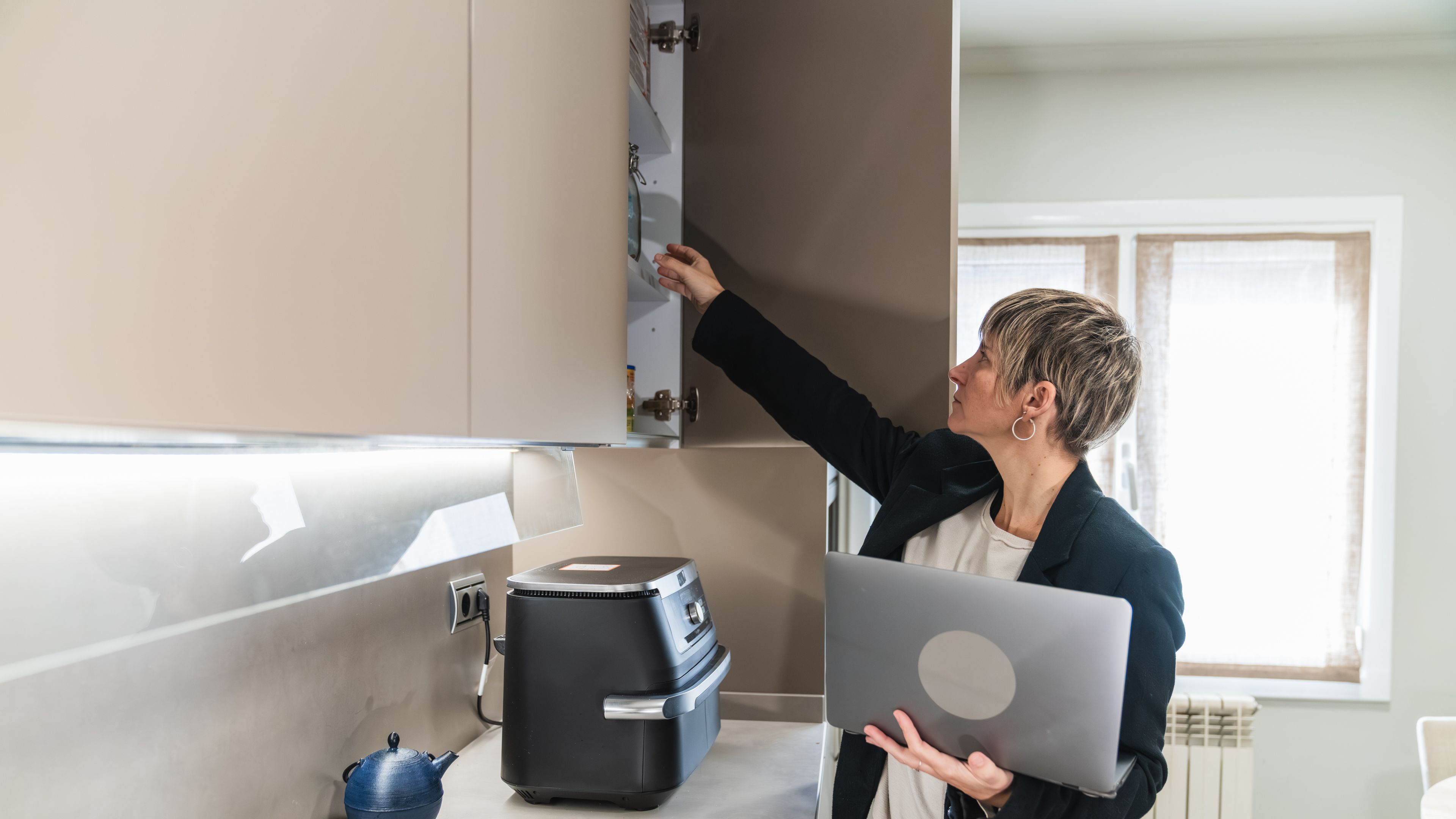 Woman holding laptop looking inside kitchen cabinet
