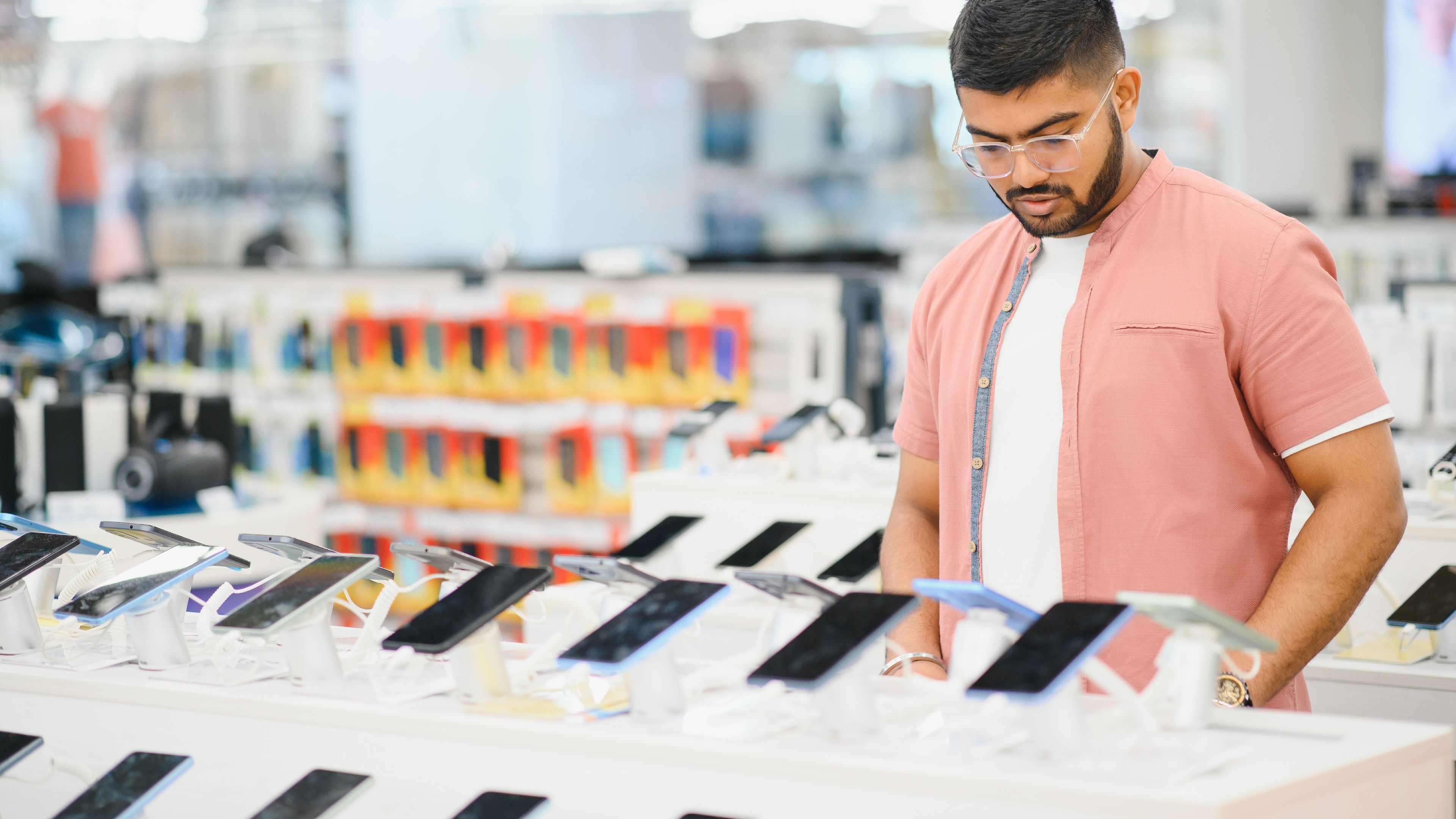 A man, dressed casually, stands at a brightly lit mobile phone store, thoughtfully comparing smartphone models 