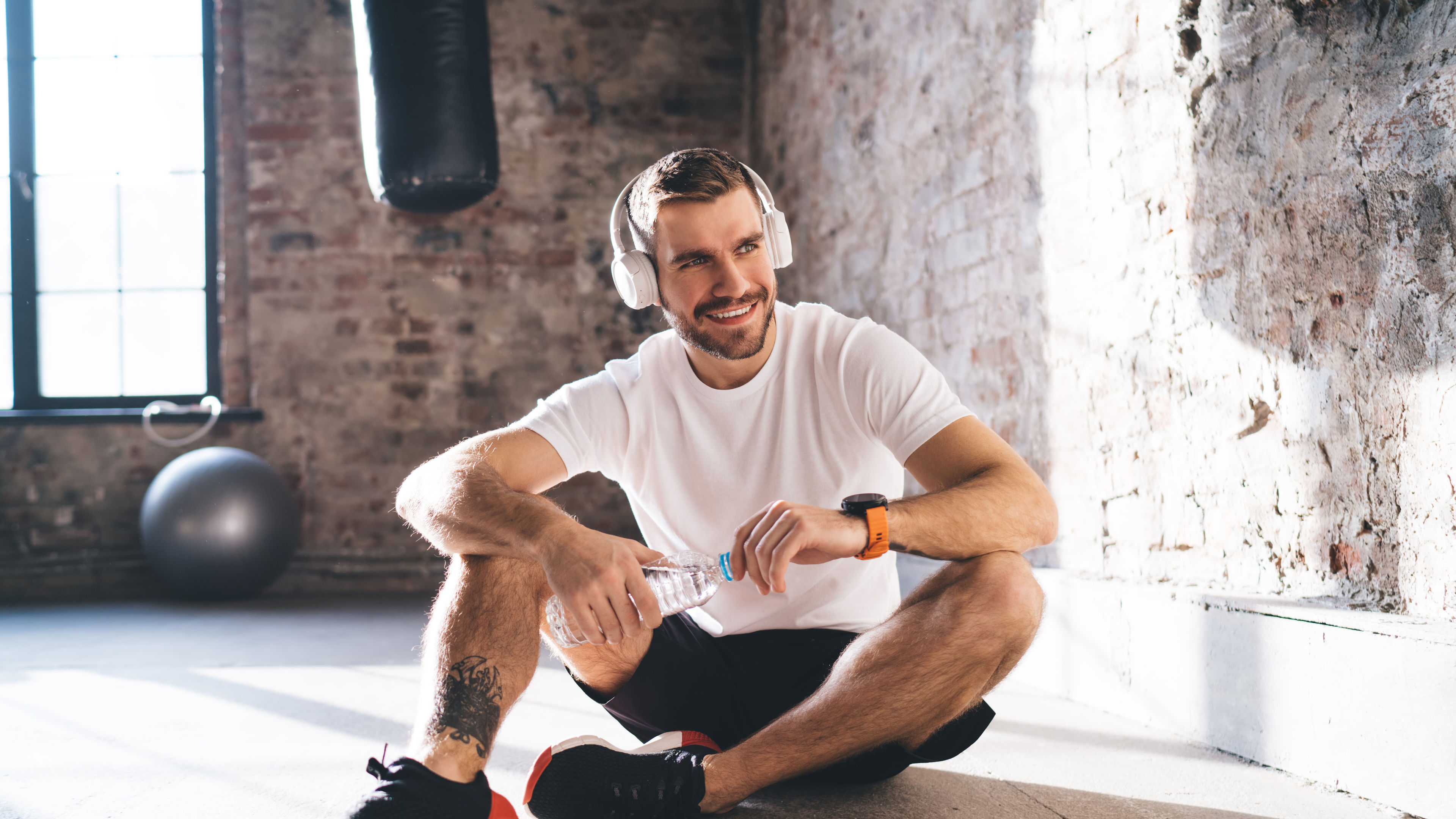 A sportsman sitting on the floor with headphones on, holding a bottle of drinking water in his hands.