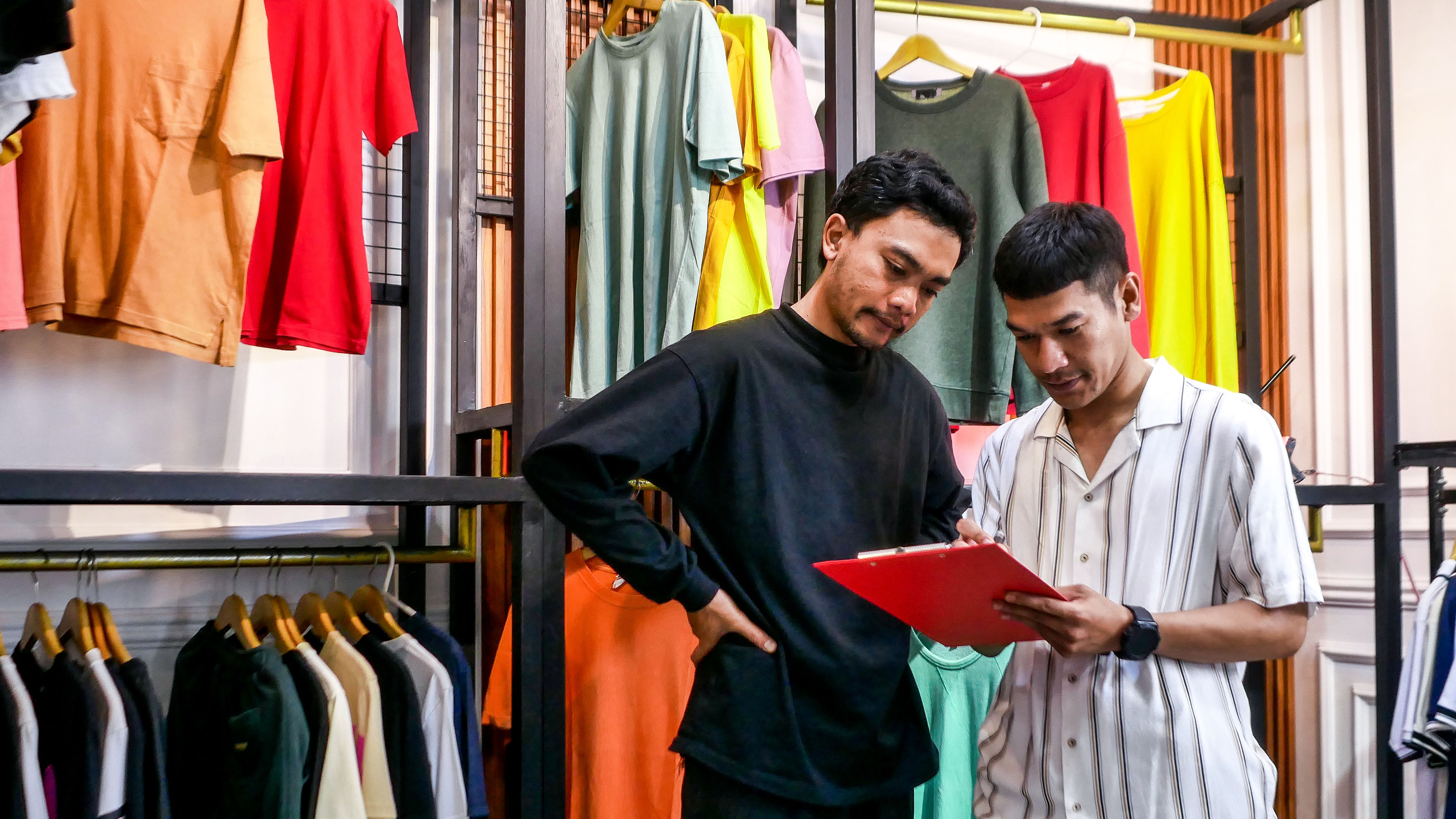 Two Young Men Collaborate Reviewing Inventory on a Clipboard Amidst Colourful Apparel Displays 

 