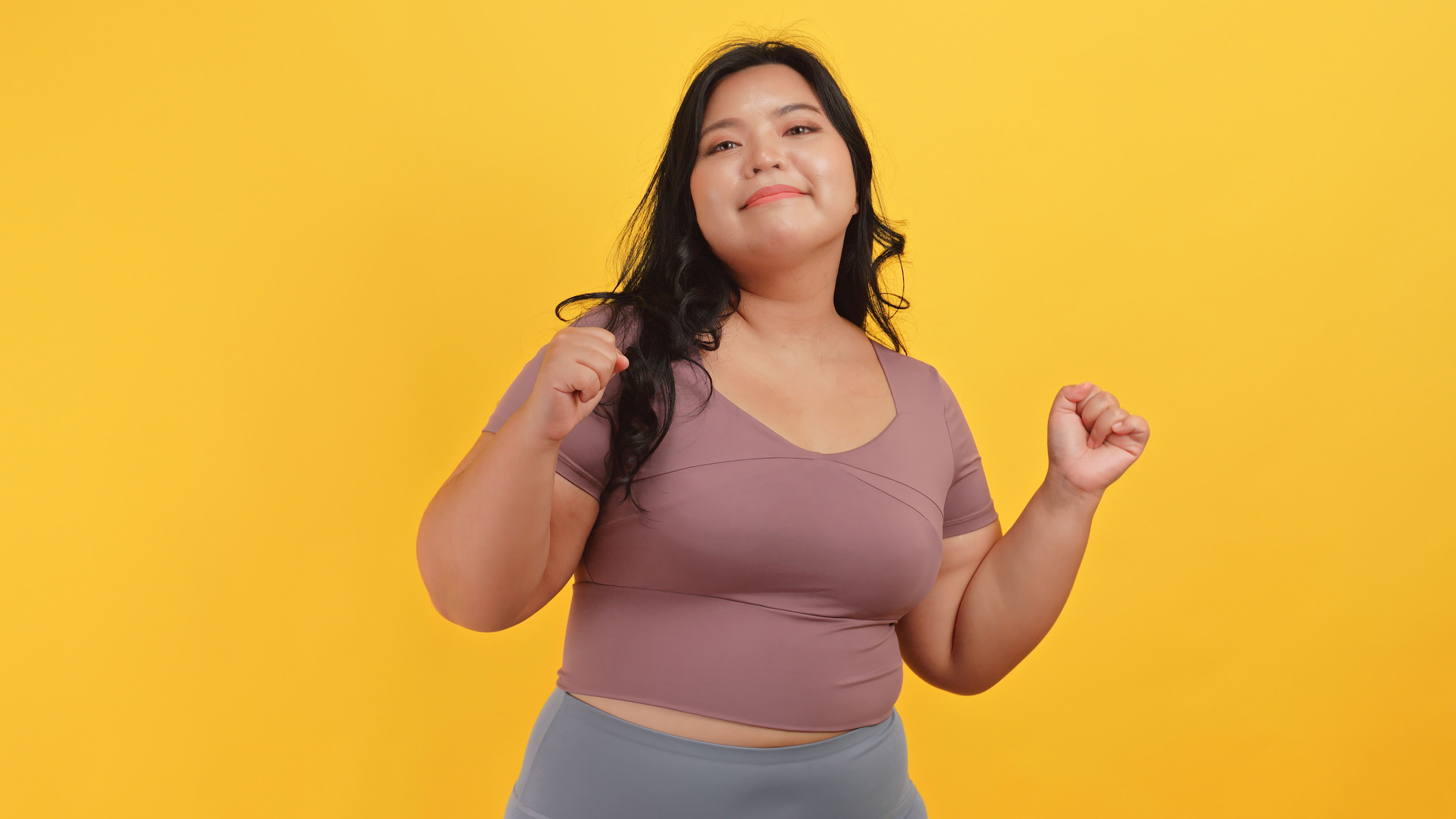 A chubby Asian woman in sportswear is having fun dancing on a yellow background. 
