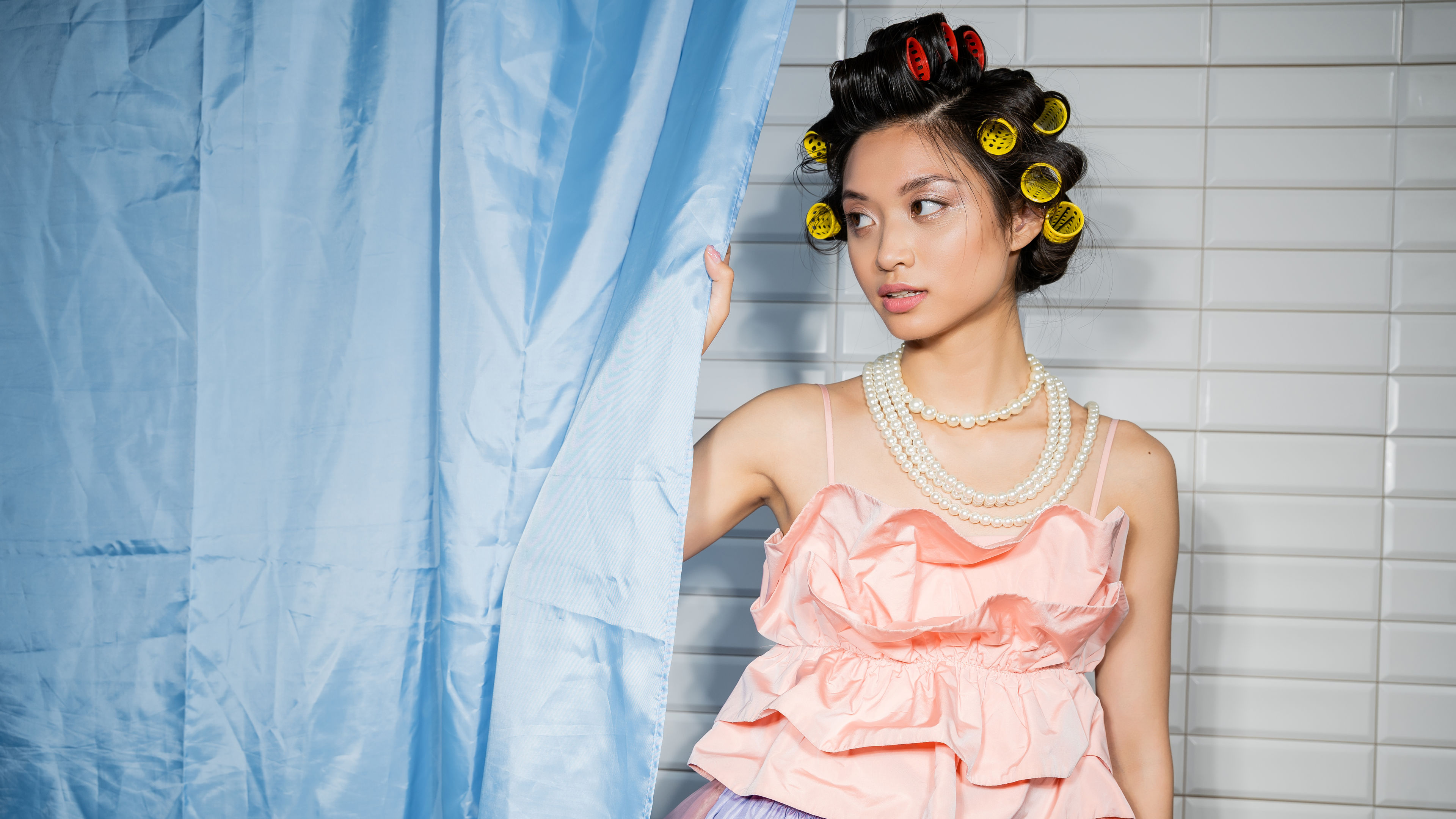 Asian young woman with hair curlers standing in pink ruffled top with pearl necklace 
