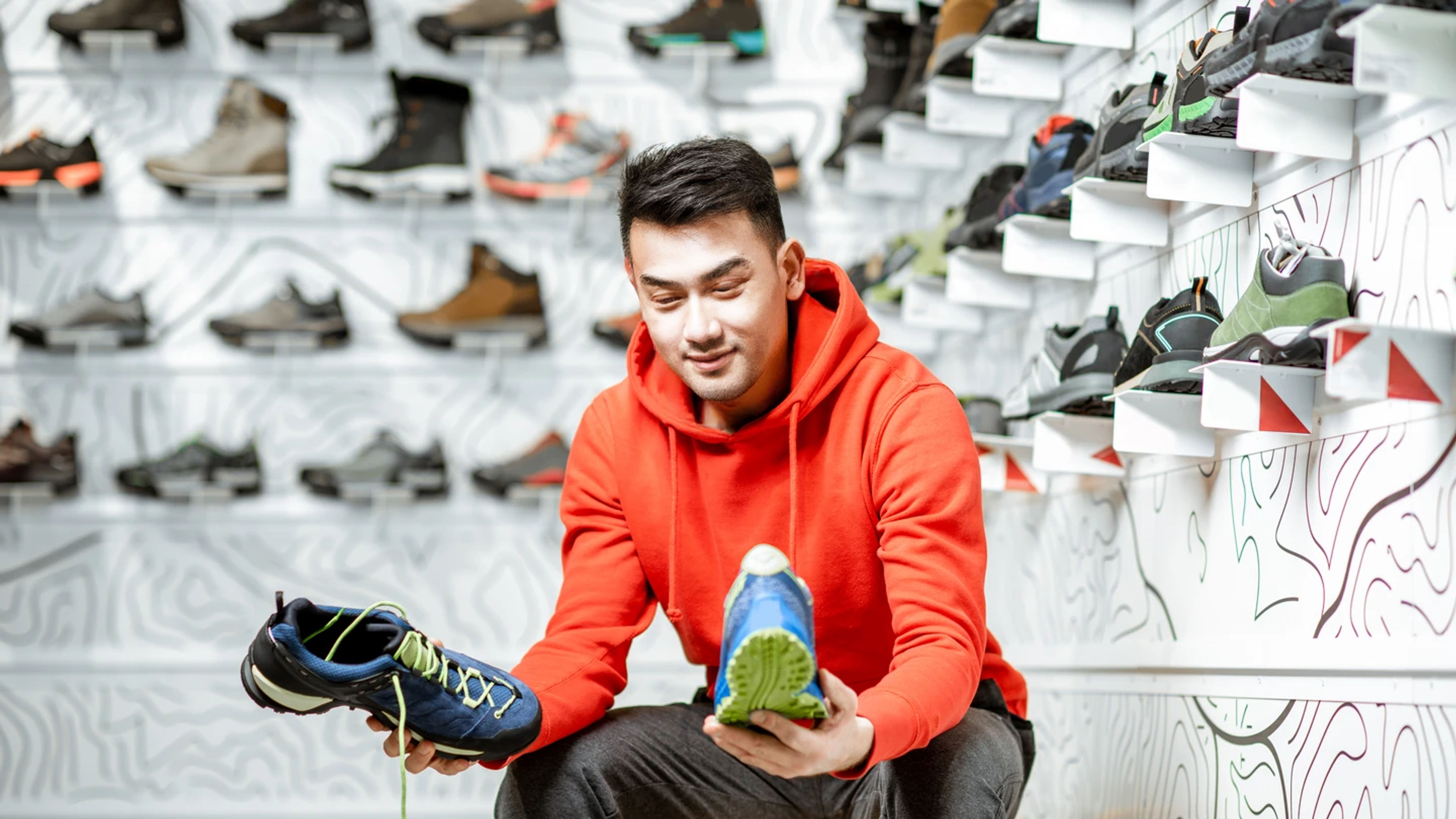 An Asian man in orange hoodie choosing hiking shoes in the shop.