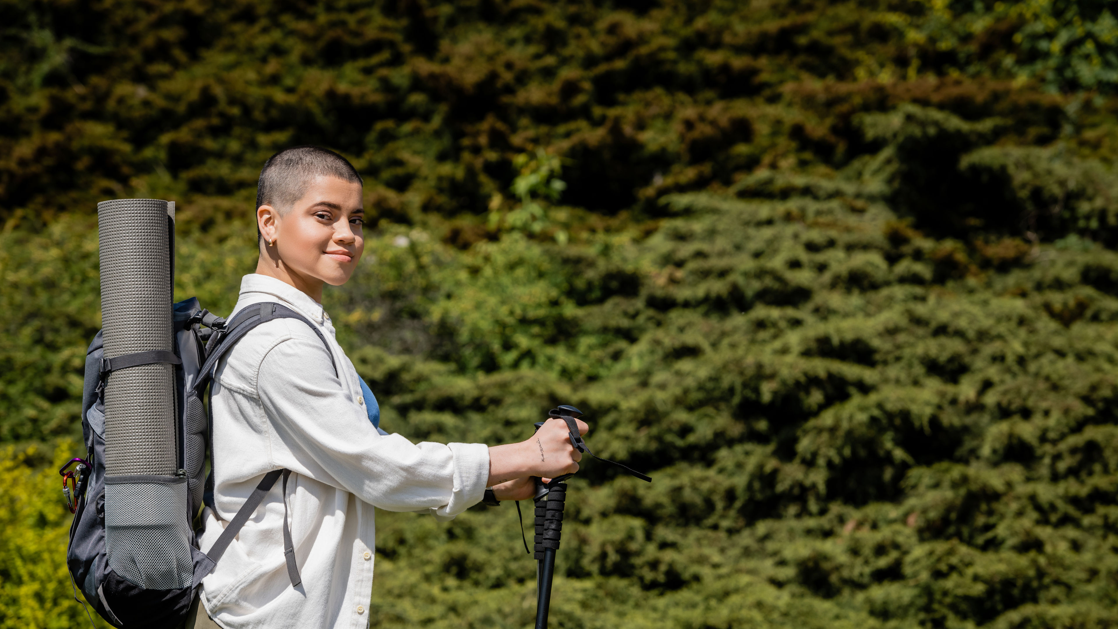 Smiling young short haired female hiker with backpack holding trekking poles and looking at camera 